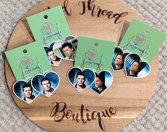 May include: Heart-shaped earrings featuring images of actors, displayed on green cards with a logo. The earrings are on a wooden display board with the words "Wild Thread Boutique" engraved on it.