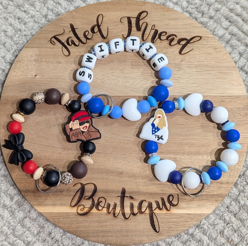 May include: Three beaded bracelets with charms and key rings are displayed on a wooden board. One bracelet spells out "SWIFTIE" with letter beads. The other bracelets have charms with a cartoon image and a woman in a dress.