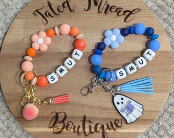 May include: Two beaded wrist keychains. One is orange and peach with a peach charm and the word "SMUT". The other is blue with a ghost charm and the word "SMUT". Both have a flower accent.