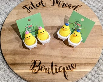 May include: Four pairs of novelty earrings featuring yellow rubber duckies in white bathtubs. Each ducky has a small hat. The earrings are displayed on green cards with the text "Fated Thread" and a logo, arranged on a wooden board.