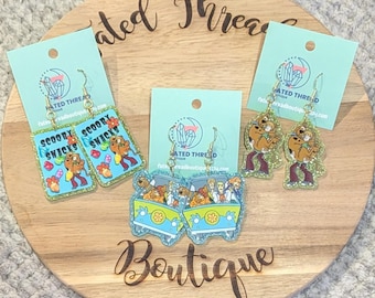 May include: Three pairs of dangle earrings featuring Scooby-Doo characters. One pair is rectangular with Scooby Snacks text, one pair shows the Mystery Machine van, and one pair features Scooby-Doo and Shaggy. All have glittery edges and gold hooks.