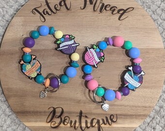 May include: Two beaded keychains with colorful beads in purple, blue, pink, and green. Each keychain features a charm with a yarn and needle design and the words "Handmade with Love". The keychains are displayed on a wooden board with the text "Tated Thread Boutique".