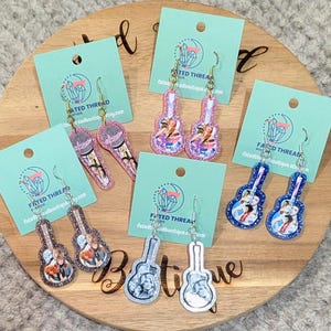 May include: Five pairs of dangle earrings on light blue display cards. One pair features microphones, while four pairs are guitar-shaped. The designs include colorful printed images of musicians, accented with glittery borders in pink, blue, and silver tones.