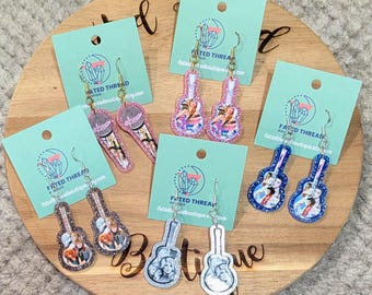 May include: Five pairs of dangle earrings on light blue display cards. One pair features microphones, while four pairs are guitar-shaped. The designs include colorful printed images of musicians, accented with glittery borders in pink, blue, and silver tones.