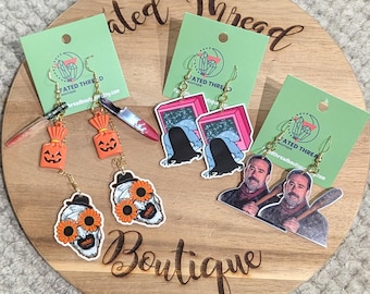 May include: A collection of Halloween-themed earrings displayed on a wooden board. The earrings feature various designs, including jack-o'-lanterns, a bloody knife, a skull with sunflowers, and characters from horror movies. The earrings are attached to green cards.