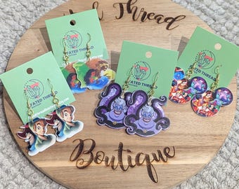 May include: Earrings featuring various cartoon characters, including Peter Pan, Ursula, and other Disney characters. The earrings are displayed on green cards and arranged on a wooden, circular display.