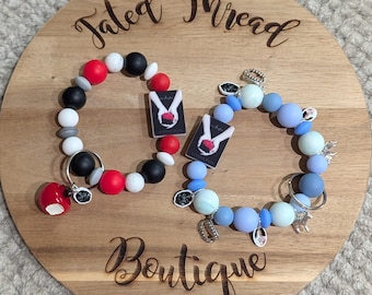 May include: Two beaded bracelets with charms, one in red, black, white, and gray, and the other in shades of blue. Each bracelet features a small book charm. The bracelets are displayed on a wooden board with the text "Tated Thread Boutique."