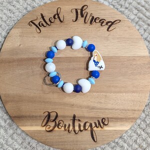 May include: A beaded keychain bracelet with white, blue, and navy beads, and a charm with a blonde woman in a blue dress. The bracelet is on a wooden round board with the words "Tated Thread Boutique" engraved on it.
