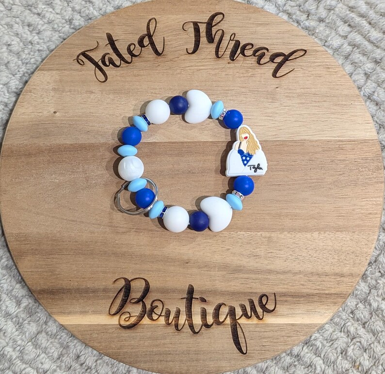 May include: A beaded keychain bracelet with white, blue, and navy beads, and a charm with a blonde woman in a blue dress. The bracelet is on a wooden round board with the words "Tated Thread Boutique" engraved on it.