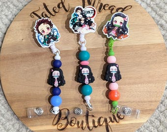 May include: Three anime-themed badge reels with character charms and colorful beads. The reels feature characters from the anime series, with a variety of bead colors including blue, pink, and green. The reels are displayed on a wooden surface with the text "Tated Thread Boutique".