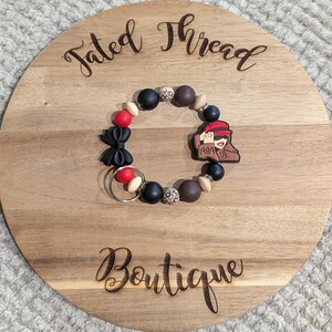 May include: A beaded bracelet with black, red, brown, and cream-colored beads. The bracelet features a black bow, a charm with a person wearing a red hat, and the number "1989". The bracelet is displayed on a wooden round board with the words "Tated Thread Boutique".