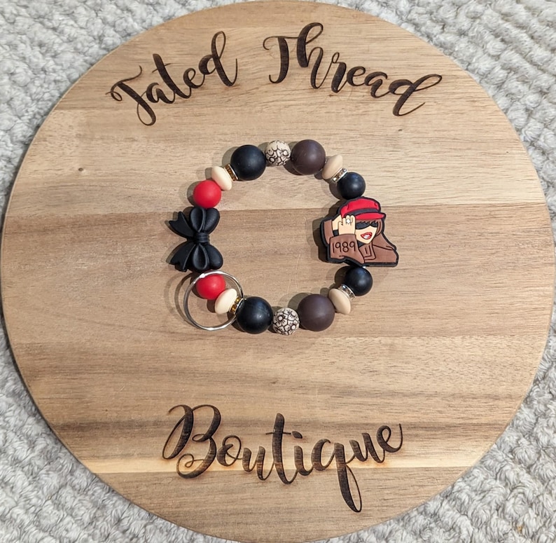 May include: A beaded bracelet with black, red, brown, and cream-colored beads. The bracelet features a black bow, a charm with a person wearing a red hat, and the number "1989". The bracelet is displayed on a wooden round board with the words "Tated Thread Boutique".