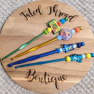 May include: Four crochet hooks with decorative tops rest on a wooden board. The hooks are green, gold, and blue, with colorful beads and charms. The board has the words "Fated Thread Boutique" engraved on it.