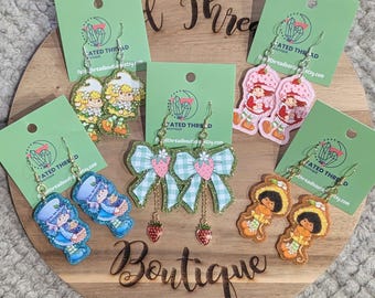 May include: A collection of colorful, glittery dangle earrings featuring cartoon characters and strawberry designs. The earrings are displayed on green cards and a wooden board with the word "Boutique".