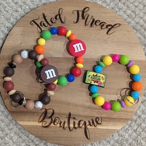 May include: Three beaded keychains with colorful beads and charms, including M&M's and Sour Patch Kids designs, are displayed on a wooden board. The board has the words "Fated Thread Boutique" engraved on it.