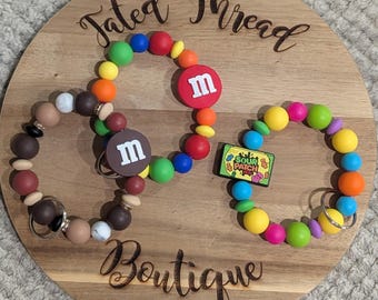 May include: Three beaded keychains with colorful beads and charms, including M&M's and Sour Patch Kids designs, are displayed on a wooden board. The board has the words "Fated Thread Boutique" engraved on it.