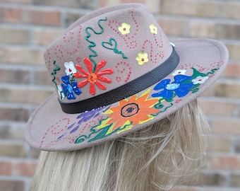 May include: A brown felt fedora hat with a black leather band. The hat is decorated with hand-painted flowers in various colors, including red, yellow, blue, orange, and green.