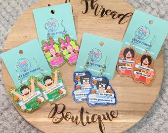 May include: Four pairs of glittery acrylic dangle earrings featuring cartoon characters from Bob's Burgers. Each pair is attached to a light blue display card with the Fated Thread Boutique logo. The designs include text bubbles with phrases like Your favorite fruity boy, Past, Your ass is grass and I'm gonna mow it, and Nonsense or MOM-sense.