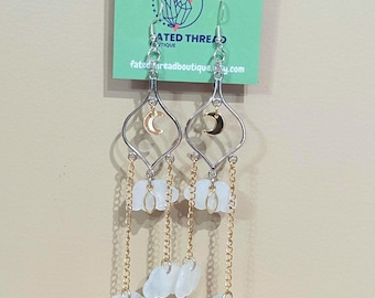 May include: Silver-toned dangle earrings with a crescent moon and cloud design. The earrings feature a gold chain and white cloud charms. The earrings are hanging from a green tag that says "FATED THREAD BOUTIQUE" and "fatedthreadboutique.com".