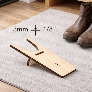 Könnte beinhalten: Ein hölzerner Stiefelknecht mit hellbraunem Finish, der das Ausziehen von Stiefeln erleichtern soll. Der Stiefelknecht liegt auf einem grauen Teppich, im Hintergrund ein Paar braune Lederstiefel. Der Stiefelknecht ist 3 mm dick.