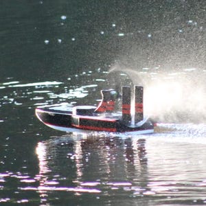 Puede incluir: Un pequeño barco modelo negro y rojo con tres chimeneas se mueve por el agua, creando un chorro de agua. El barco tiene una base blanca y se refleja en el agua.