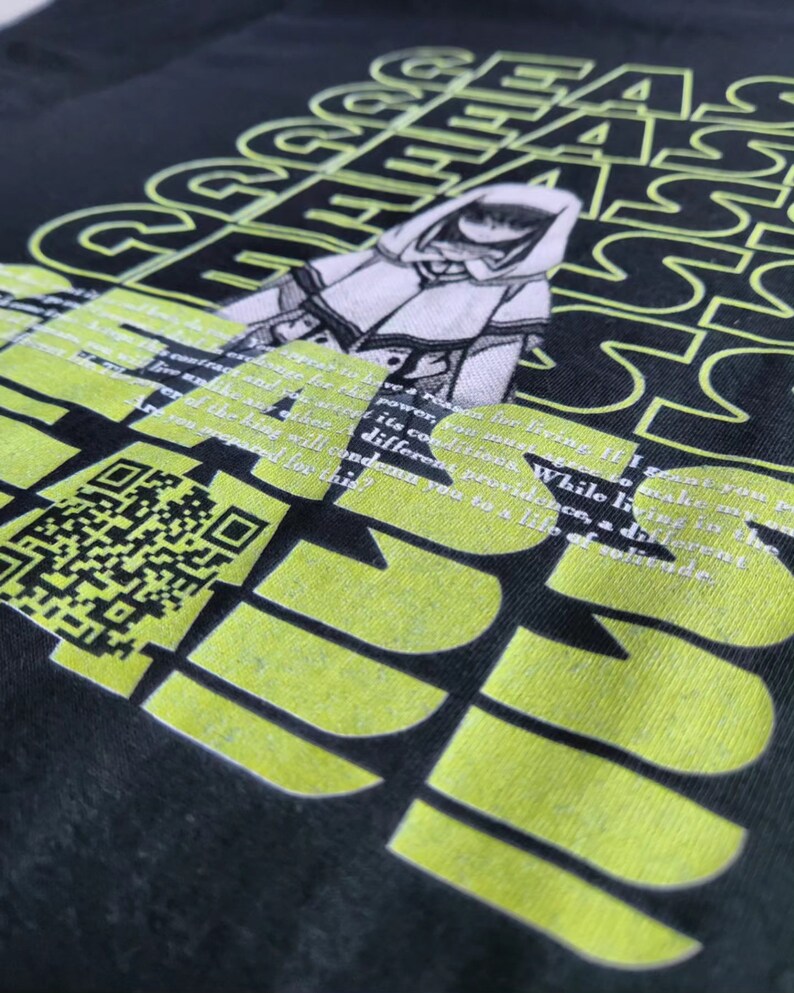 Op de afbeelding: Zwart T-shirt met een limoengroen grafisch ontwerp. Het ontwerp bevat een gestileerde illustratie van een figuur met een capuchon, tekst en een QR-code. De tekst bevat de woorden "macht", "anders" en "veroordelen".