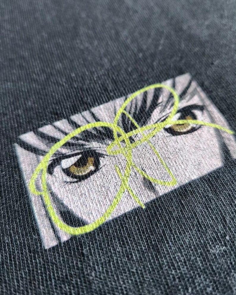 Op de afbeelding: Close-up van een donkergrijze stof met een geborduurd ontwerp van ogen in anime-stijl. De ogen zijn bruin met witte highlights, tegen een grijze en witte achtergrond. Een neon groen lijnkunstontwerp bedekt de ogen.