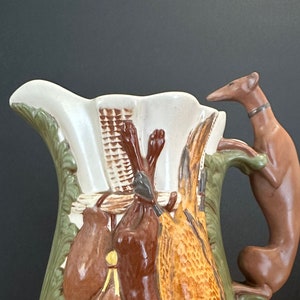 May include: A decorative ceramic pitcher with a brown dog-shaped handle. The pitcher features relief artwork of game birds, a basket, and foliage in shades of brown, green, and cream. The pitcher is approximately 10 inches tall.