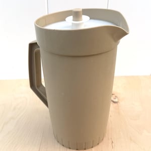 May include: A tan plastic pitcher with a white lid and handle. The pitcher has a spout for pouring and is designed for holding liquids. The pitcher is sitting on a light wood surface.