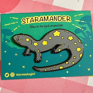 May include: A green enamel pin featuring a gray salamander with yellow stars. The pin reads "Staramander" and "Glow in the dark enamel pin".