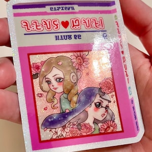 Puede incluir: Una pegatina rosa y blanca con una ilustraci&oacute;n de dibujos animados de dos chicas con flores. La pegatina tiene el texto "2 girls &hearts; 1 story" y "99 bottle" en rosa.