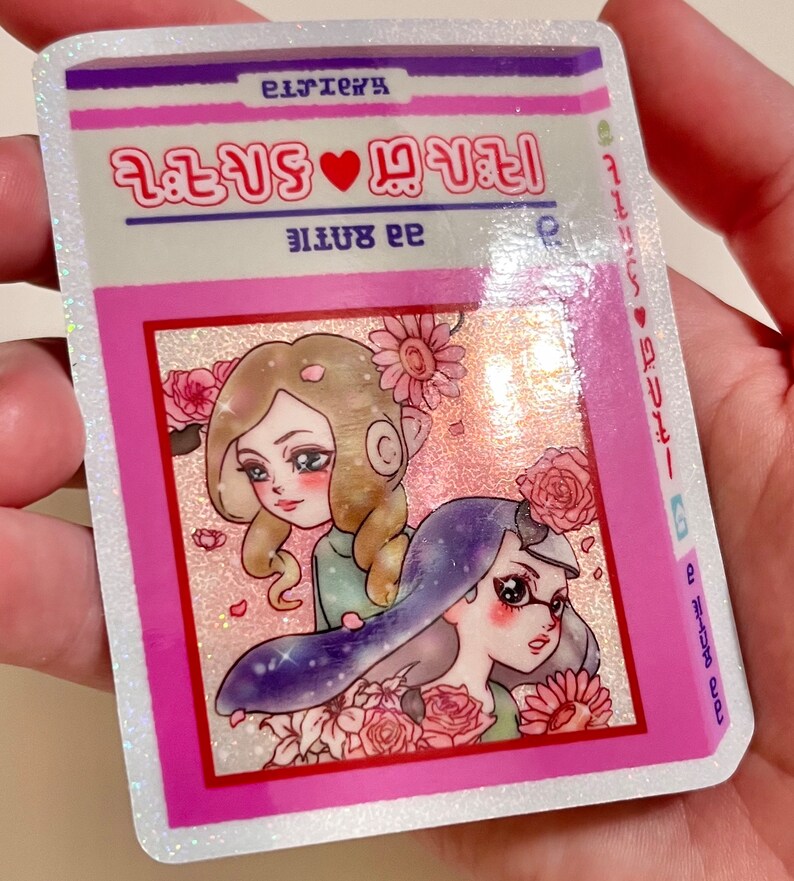 Puede incluir: Una pegatina rosa y blanca con una ilustraci&oacute;n de dibujos animados de dos chicas con flores. La pegatina tiene el texto "2 girls &hearts; 1 story" y "99 bottle" en rosa.