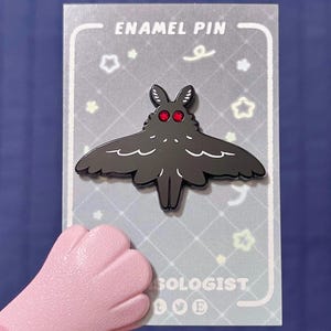 Pode incluir: Um pin de esmalte preto em forma de traça com olhos vermelhos, exibido em um cartão cinza com as palavras "ENAMEL PIN". O pin tem detalhes brancos e é segurado por uma mão rosa. O cartão tem desenhos de estrelas e redemoinhos.