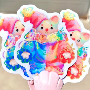 May include: A stack of colorful stickers featuring a whimsical cartoon bear dressed in a clown-like outfit. The bear has a white face, pink ears, and is wearing a hat. The stickers have a rainbow color scheme with glitter accents.