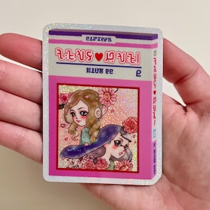 Puede incluir: Una pegatina rosa y blanca con un acabado brillante. La pegatina presenta una ilustraci&oacute;n de dibujos animados de dos chicas con flores. El texto en la pegatina dice "EIJITENA" y "22 AUG".