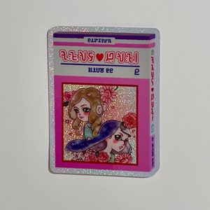 Puede incluir: Una pegatina rosa y blanca con un acabado brillante. La pegatina presenta una ilustraci&oacute;n de dos chicas con flores y el texto "2 girls &hearts; 2 girls" en un coraz&oacute;n rosa. El texto "2 girls" se repite en el lado derecho de la pegatina.
