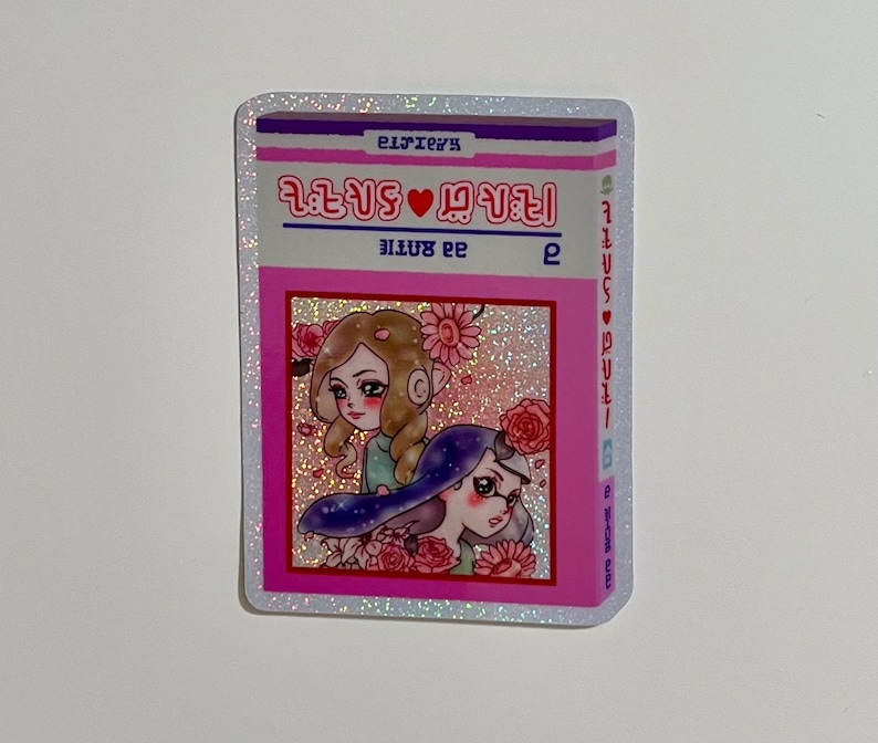 Puede incluir: Una pegatina rosa y blanca con un acabado brillante. La pegatina presenta una ilustraci&oacute;n de dos chicas con flores y el texto "2 girls &hearts; 2 girls" en un coraz&oacute;n rosa. El texto "2 girls" se repite en el lado derecho de la pegatina.