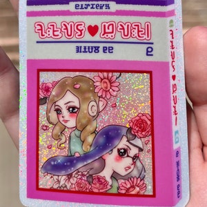 Puede incluir: Una pegatina rosa y blanca con fondo brillante. La pegatina presenta dos chicas de dibujos animados con flores y el texto "2 girls &hearts; 1 planet" en un coraz&oacute;n rosa. El texto "e 22118" est&aacute; en el lado derecho de la pegatina.