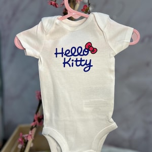 Pu&ograve; includere: Un body bianco per neonati con la scritta "Hello Kitty" in blu e un fiocco rosso. Il body &egrave; appeso a una gruccia rosa, con fiori rosa sullo sfondo.