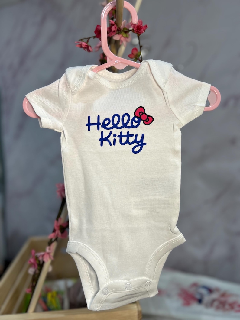 Pu&ograve; includere: Un body bianco per neonati con la scritta "Hello Kitty" in blu e un fiocco rosso. Il body &egrave; appeso a una gruccia rosa, con fiori rosa sullo sfondo.