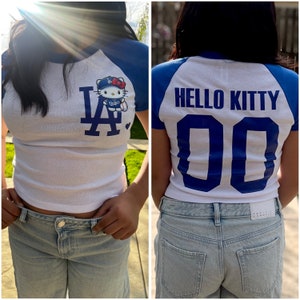 Pu&ograve; includere: T-shirt bianco e blu in stile baseball con il personaggio di Hello Kitty che indossa un berretto da baseball e un'uniforme dei Dodgers. La parte anteriore della maglietta mostra il logo dei Dodgers. La parte posteriore della maglietta dice "HELLO KITTY" e il numero "00".