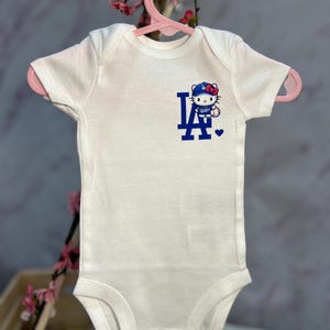 Pu&ograve; includere: Body per neonati bianco con maniche corte, con logo blu "LA" e un personaggio di Hello Kitty con un berretto da baseball. Il body &egrave; appeso a un gancio rosa.