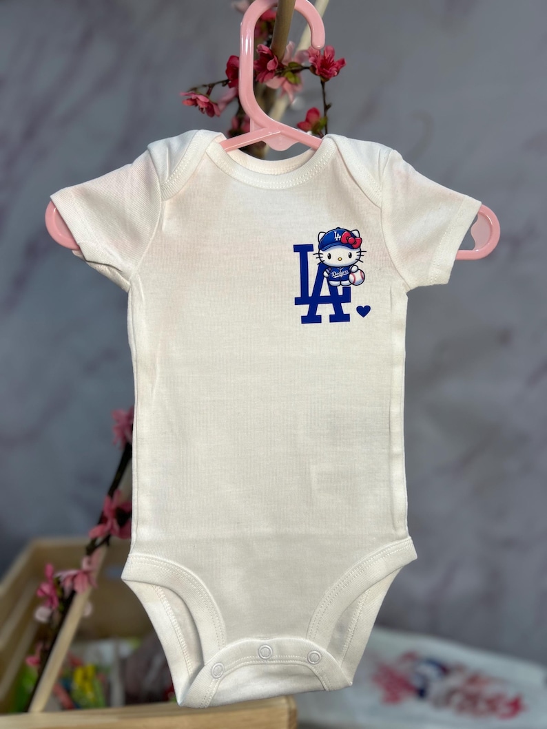 Pu&ograve; includere: Body per neonati bianco con maniche corte, con logo blu "LA" e un personaggio di Hello Kitty con un berretto da baseball. Il body &egrave; appeso a un gancio rosa.