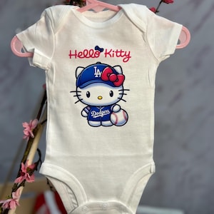 Pu&ograve; includere: Body da bambino bianco con una grafica di Hello Kitty che indossa un berretto da baseball blu con le lettere "LA" e un fiocco rosso. La grafica include anche una palla da baseball e la scritta "Dodgers".