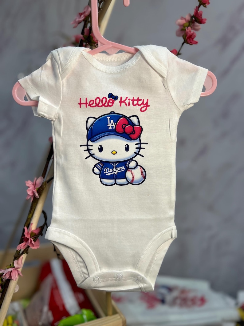 Pu&ograve; includere: Body da bambino bianco con una grafica di Hello Kitty che indossa un berretto da baseball blu con le lettere "LA" e un fiocco rosso. La grafica include anche una palla da baseball e la scritta "Dodgers".