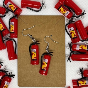Puede incluir: Pendientes rojos con forma de extintor con ganchos plateados. Los pendientes en miniatura tienen asas negras y etiquetas amarillas y negras. El fondo presenta más réplicas de extintores, creando un tema lúdico.