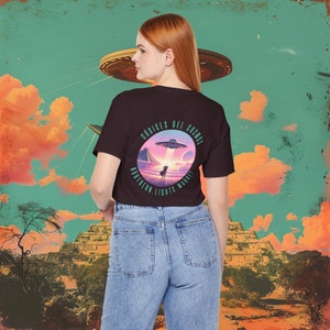 May include: A black t-shirt with a circular graphic on the back featuring a UFO, a person, and the text "CODIGES DEL COSMOS" and "NORTHERN LIGHTS MARKET". The background includes a pyramid and a colorful sky.