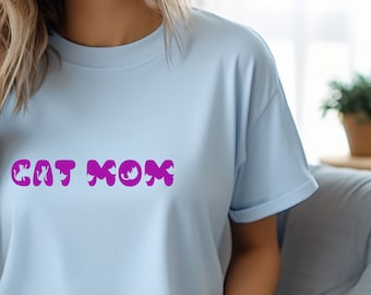 May include: A light blue t-shirt with the text "CAT MOM" in a vibrant purple color. The text has a playful, rounded font. The shirt is made of a soft material and has short sleeves.