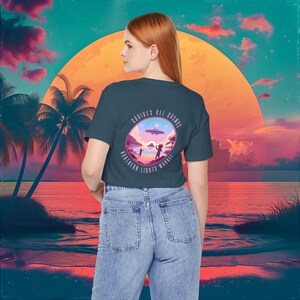 May include: A dark teal t-shirt with a pink and purple graphic of a beach scene with two figures and a flying saucer. The graphic also includes the text "CODICES DEL COSMOS NORTHERN LIGHTS MARKET".