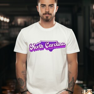 Może przedstawiać: Biała koszulka z fioletowym obrysem stanu Karolina P&oacute;łnocna i tekstem "North Carolina" w retro czcionce.